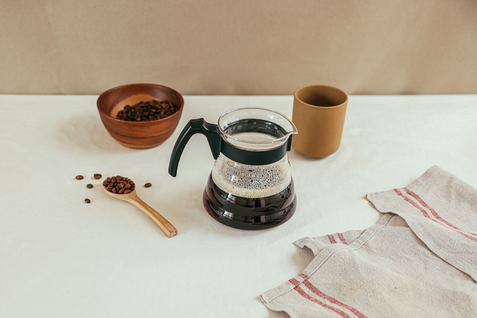 Glass pot of freshly brewed coffee with a wooden scoop of beans and a cup on a white table, the payoff of learning how to brew coffee in a pot