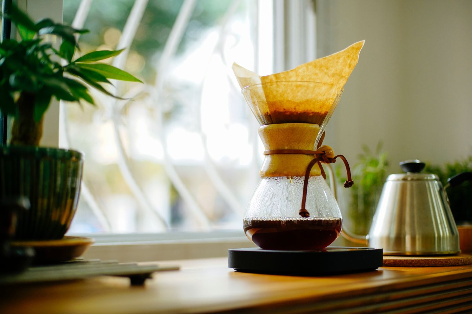 How to brew coffee at home: a glass hourglass pour-over brewer with a paper filter on a scale beside a kettle by a sunny window
